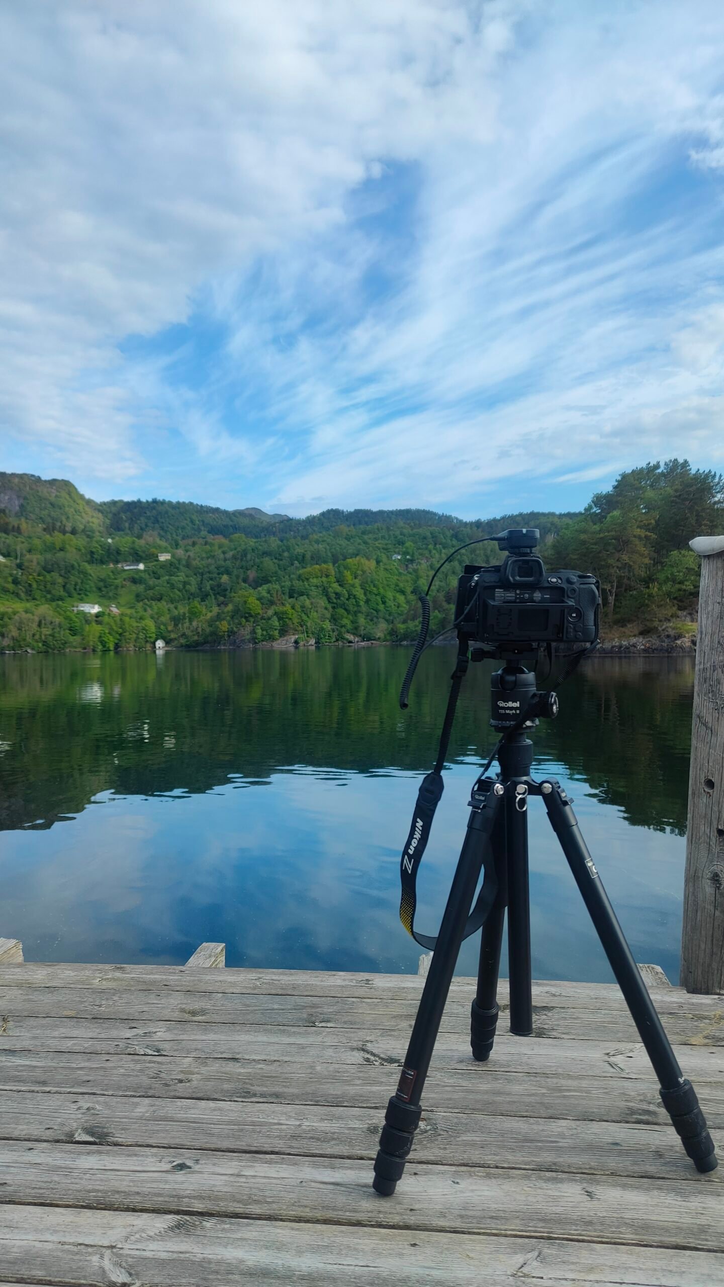 Un appareil photo Nikon installé sur un trépied. Une télécommande de déclenchement à distance est connecté à l'appareil photo. L'appareil photo fait une pause longue d'un paysage norvégien avec un lac, une forêt et un grand ciel bleue et nuageux