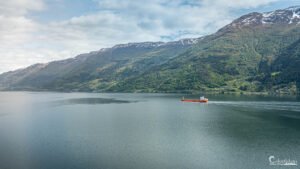 Bateau cargo rouge naviguant paisiblement sur le Hardangerfjord, entouré par des montagnes verdoyantes et une eau sereine.