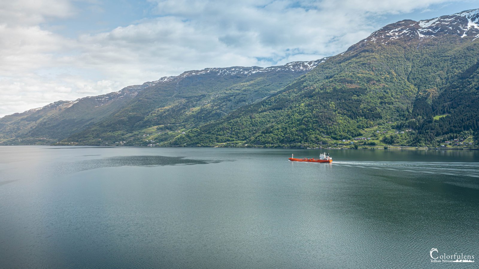 Bateau cargo rouge naviguant paisiblement sur le Hardangerfjord, entouré par des montagnes verdoyantes et une eau sereine.