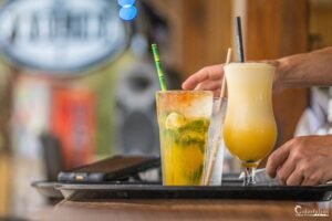 Deux cocktails tropicaux, un mojito aux feuilles de menthe et tranches de citron, et un smoothie mangue, posés sur un plateau en bois noir avec un arrière-plan flou, incarnent une pause estivale et rafraîchissante.