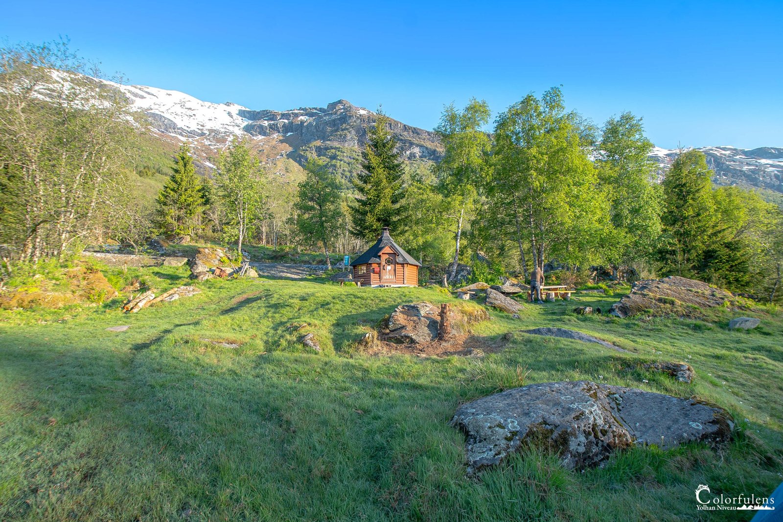 Image pittoresque d'une cabane en bois entourée de montagnes enneigées et d'une végétation luxuriante, idéale pour l'évasion et la reconnexion avec la nature.