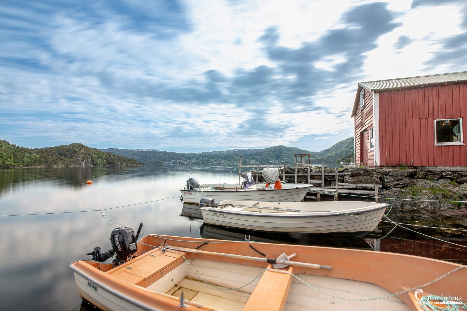 Image d'une cabane de pêcheur rouge traditionnelle au bord d'un lac norvégien avec bateaux et reflets majestueux sur l'eau calme.