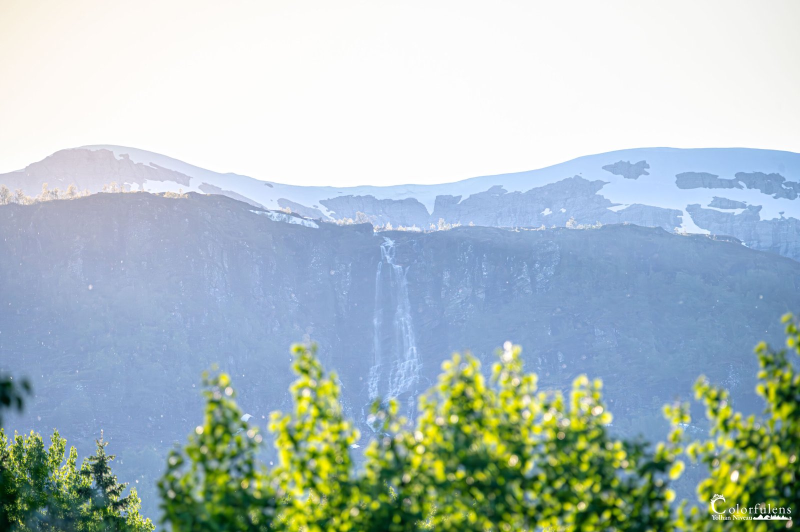 À l'aube, une cascade en montagne se déploie majestueusement, baignant la végétation verdoyante d'une lumière douce et rosée, offrant un spectacle de la nature à la quiétude matinale.