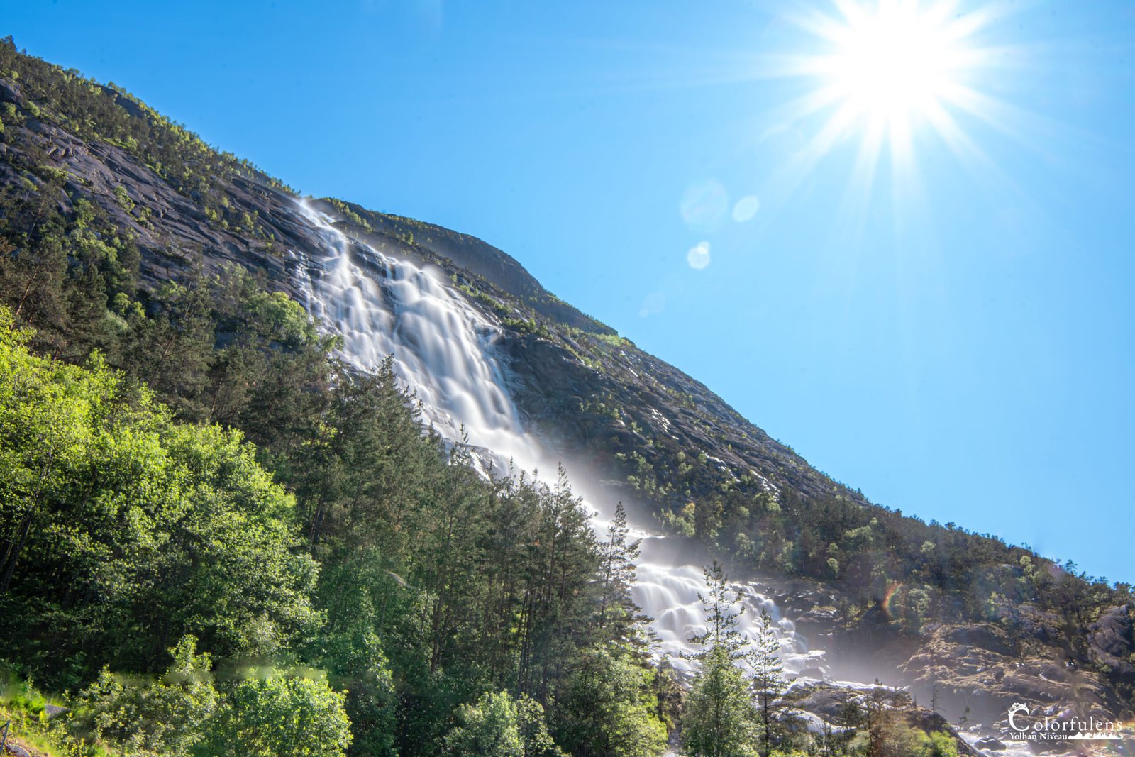 Magnifique cascade jaillissant d'une montagne sous un ciel estival, éclairée par le soleil, entourée de verdure et de roches polies par l'érosion.