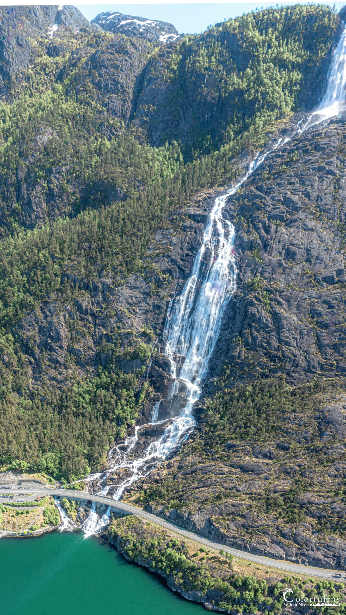 Cascade impressionnante dévalant une montagne verdoyante, entourée de nature et éblouie par les rayons du soleil, avec un pont surplombant l'eau claire.