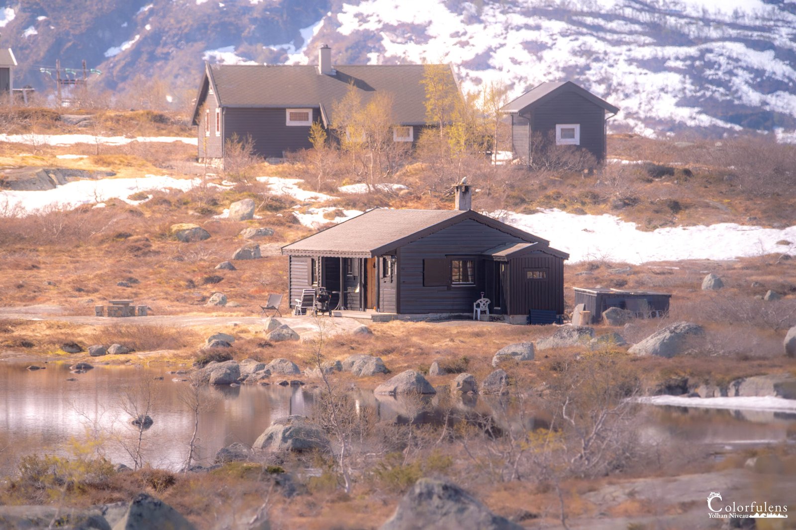 Chalet en bois au bord d'un lac de montagne, entouré de neige, rochers et végétation printanière, un havre de tranquillité.