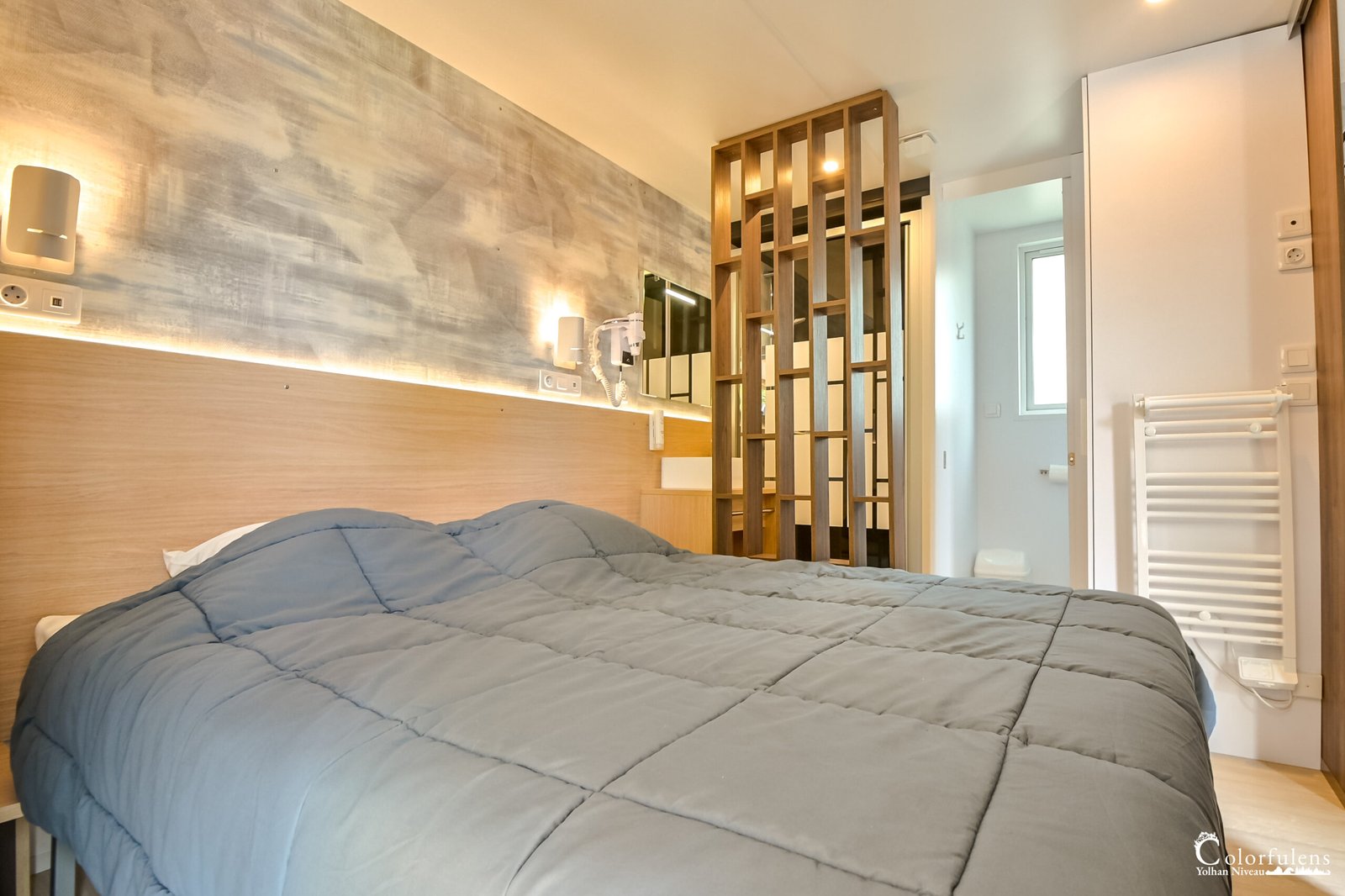 Chambre d'hôtel moderne au design intérieur épuré, avec un grand lit confortable sous une couette bleu-gris, et des détails artistiques comme le mur texturé et une séparation en bois ajouré.