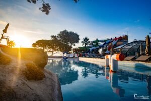 Coucher de soleil sur un complexe aquatique avec piscine, toboggans et végétation méditerranéenne, reflétant une ambiance de vacances et de détente.