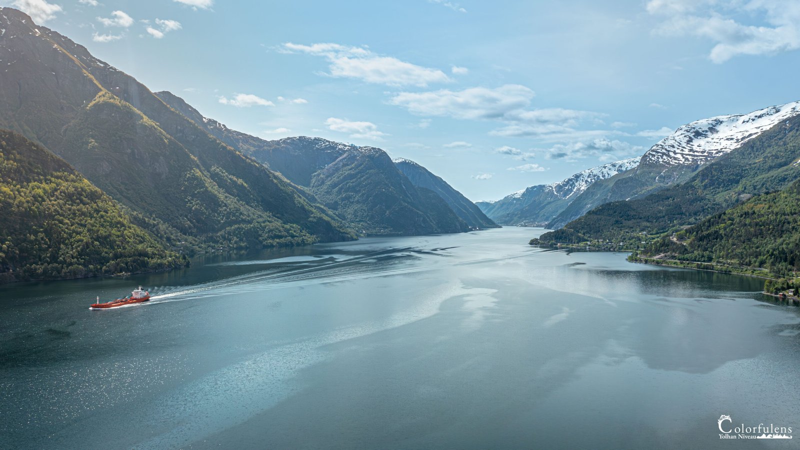 Un bateau rouge navigue paisiblement sur les eaux calmes d'un fjord norvégien, entouré de majestueuses montagnes enneigées, refletant la beauté sauvage et sereine de la nature nordique.