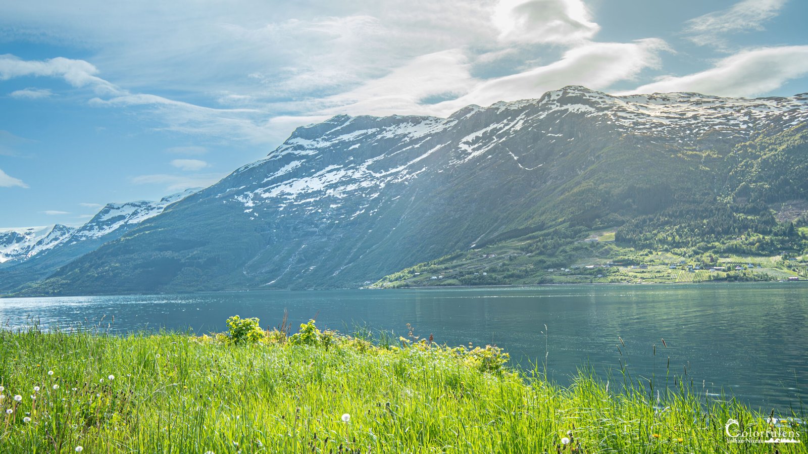 Fjord norvégien avec montagnes enneigées se reflétant dans des eaux claires sous la lumière estivale, capturant l'harmonie entre la nature et la présence humaine.
