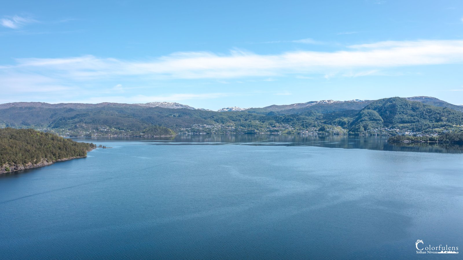 Paysage serein du fjord Samnanger, où les montagnes enneigées se reflètent dans une eau paisible et bleue, sous un ciel clair ensoleillé.