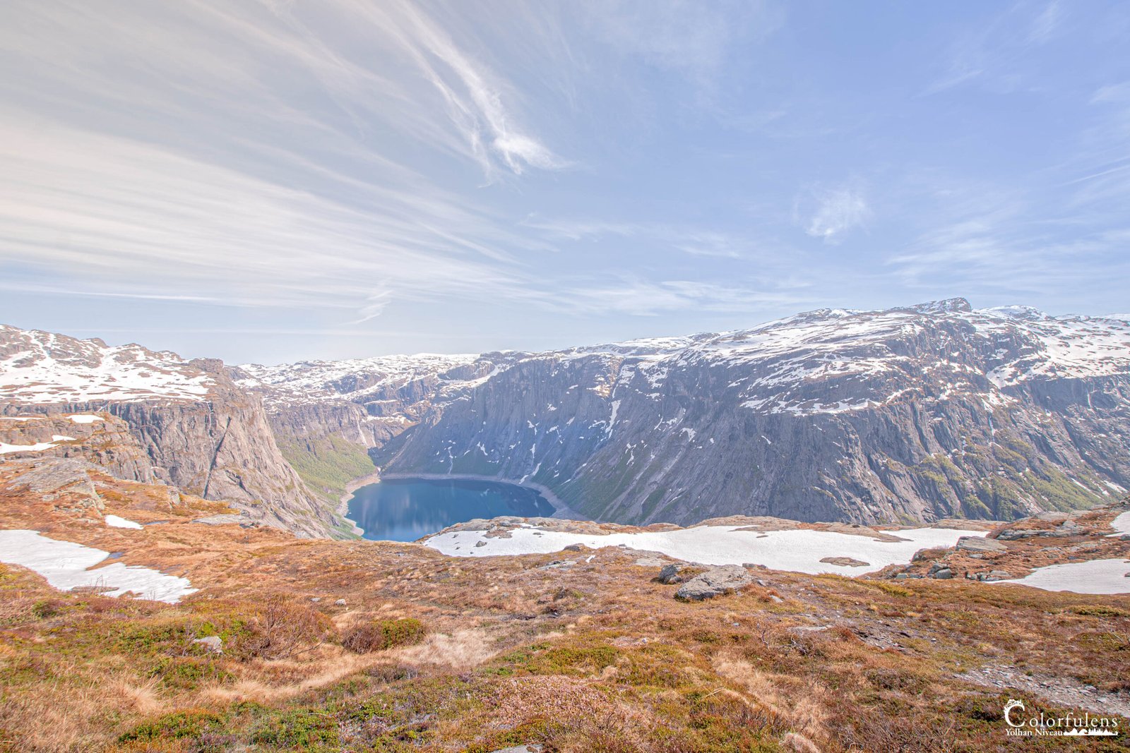 Photo panoramique d'un fjord norvégien avec un lac au centre, entouré de montagnes enneigées sous un ciel bleu clair, parfait pour les amoureux de la nature sauvage.