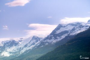 Photo de majestueuses montagnes enneigées surplombant une forêt verdoyante sous un ciel bleu parsemé de nuages, capturant la tranquillité et la beauté naturelle intacte.
