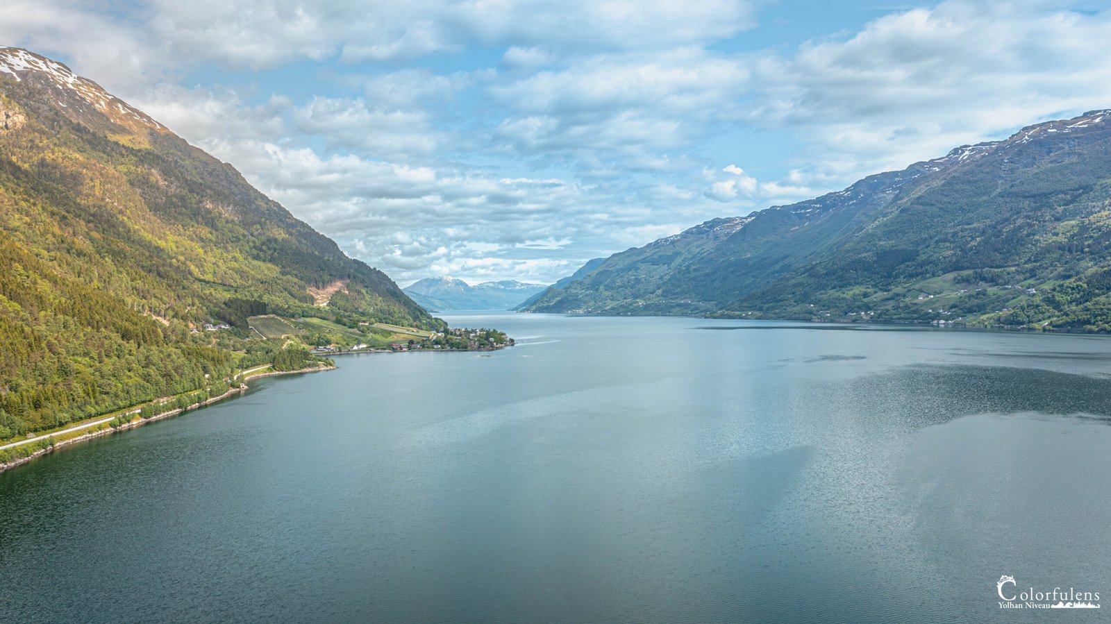 Vue aérienne du Hardangerfjord en Norvège, capturant sa majesté entre montagnes escarpées et eaux miroir, sous un ciel nuageux.