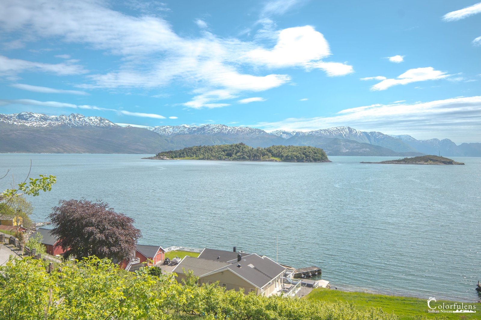 Magnifique vue d'un fjord norvégien entouré de montagnes enneigées, ciel bleu, et touches de verdure printanière, reflétant l'harmonie entre la nature sauvage et les habitations.