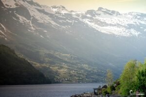 Vue sereine du fjord de Hardanger en Norvège avec des montagnes enneigées se reflétant dans une eau calme, rendant hommage à la beauté naturelle nordique.