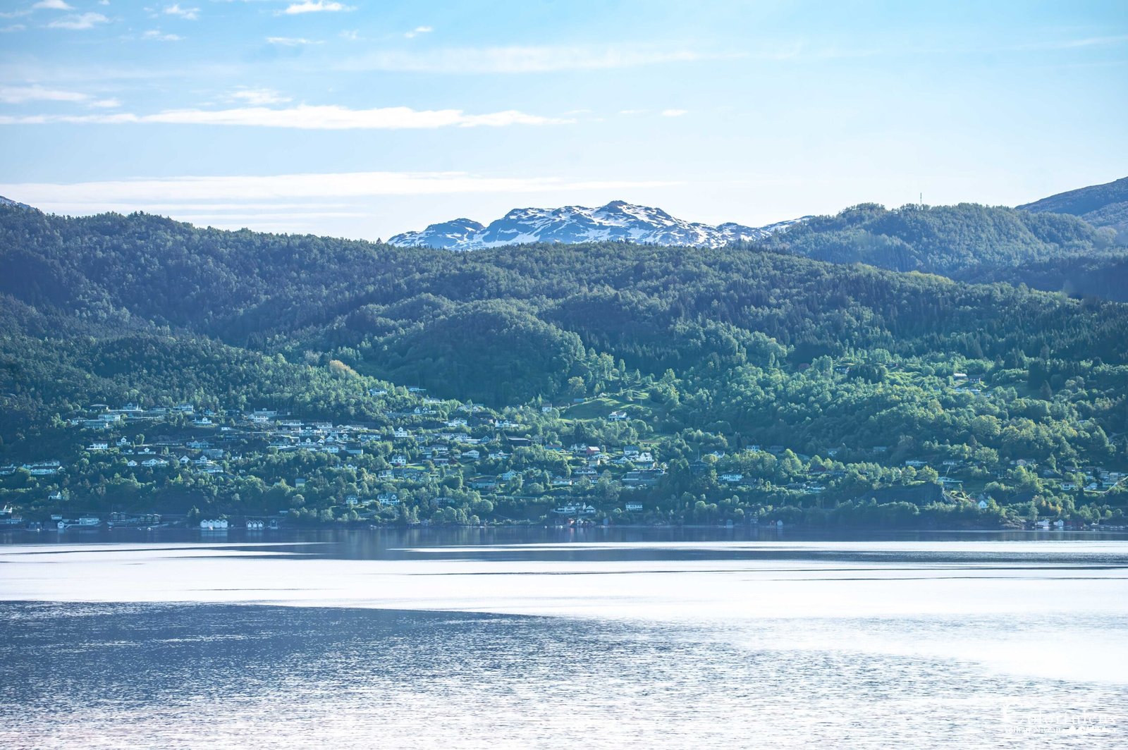Cette image montre un paysage serein de Norvège avec un lac tranquille, entouré de montagnes enneigées et de forêts verdoyantes.
