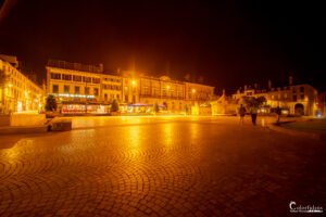 Place de ville illuminée par la lumière chaude de la nuit, avec un couple se promenant et entouré de bâtiments historiques.