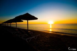 Scène sereine d'un coucher de soleil sur une plage déserte, avec des transats et parasols en silhouette offrant un spectacle pittoresque au crépuscule.