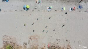 Vue aérienne d'une plage estivale animée avec parasols multicolores, serviettes, et baigneurs formant une mosaïque vibrante sous le soleil.