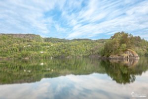 Reflet des arbres et du ciel sur un lac paisible entouré de verdure et de maisons sur une colline verdoyante.