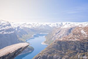 Vue aérienne depuis un rocher en Scandinavie, montrant un lac entouré de montagnes enneigées et de forêts.