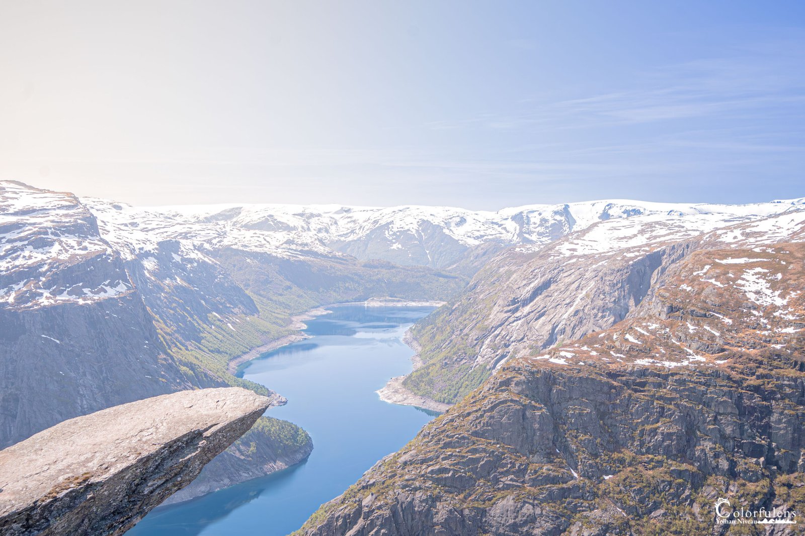 Vue aérienne depuis un rocher en Scandinavie, montrant un lac entouré de montagnes enneigées et de forêts.