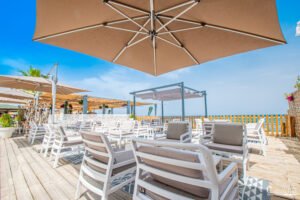 Terrasse ensoleillée avec mobilier blanc, vue sur la mer, sous une pergola de bois, entourée de plantes vibrantes et parasols.