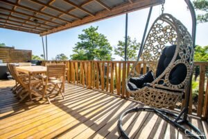 Terrasse en bois avec chaise suspendue en osier, fauteuil confortable et espace convivial pour détente ensoleillée.