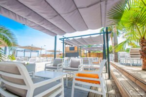 Terrasse ensoleillée avec fauteuils confortables, palmiers, et vue sur la mer, idéale pour la détente estivale en Corse.