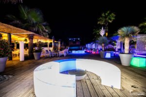 Terrasse de nuit dans un resort illuminée par des lumières LED colorées, ambiance exotique avec palmiers et mobilier moderne.