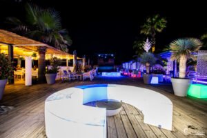 Terrasse contemporaine illuminée la nuit, proche de la plage, avec palmiers et meubles design.