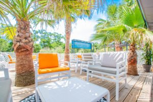 Chic et détente sur une terrasse ensoleillée de Corse, entourée de palmiers et mise en valeur par des coussins orange sur un mobilier blanc moderne.