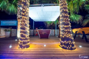 Terrasse illuminée la nuit, palmiers avec guirlandes lumineuses, ambiance de détente et convivialité sous un ciel étoilé.