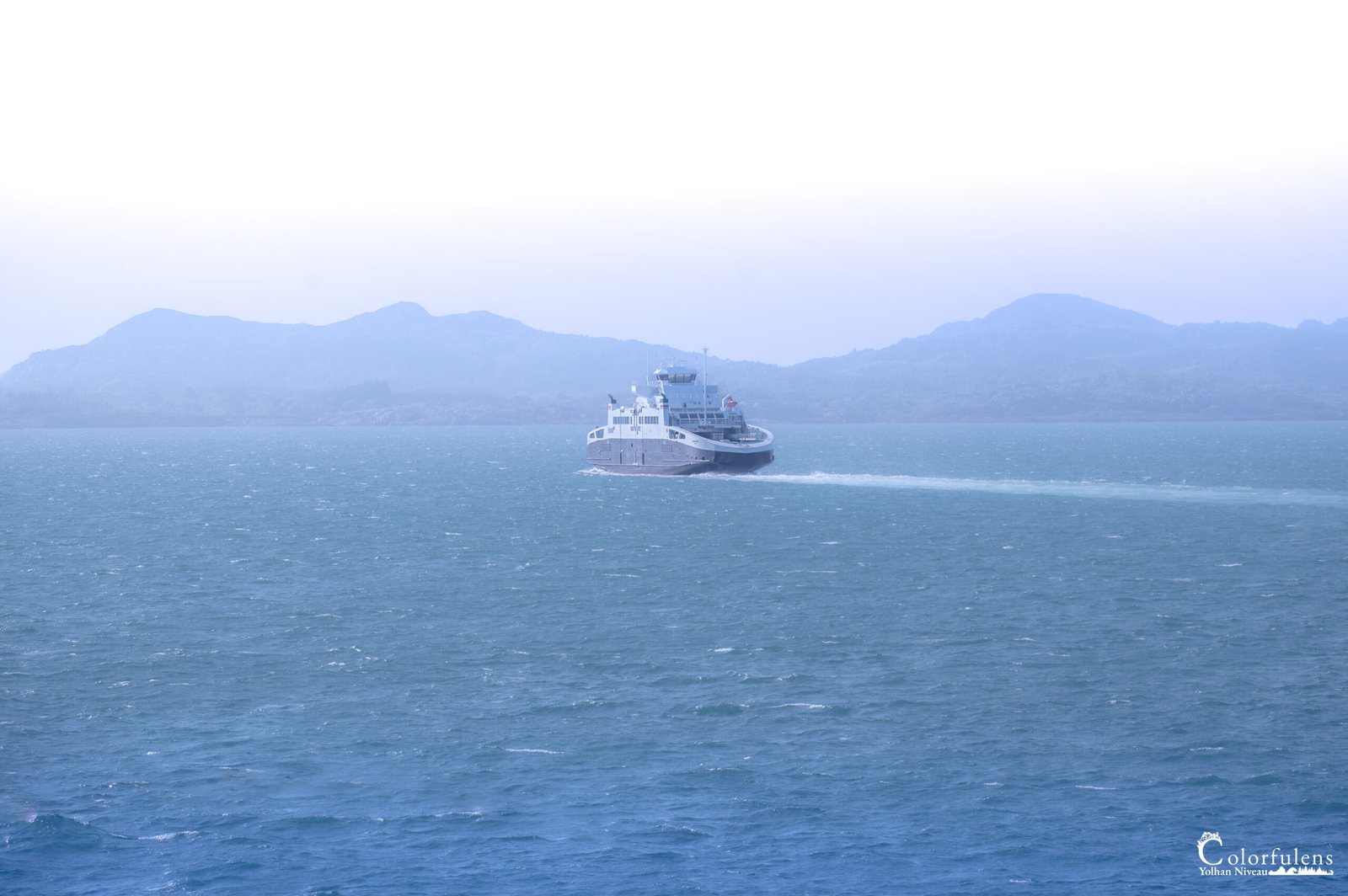 Le ferry avance paisiblement vers une montagne enveloppée de brume, offrant un voyage serein sur une mer calme, unissant mer et ciel dans une harmonie naturelle.