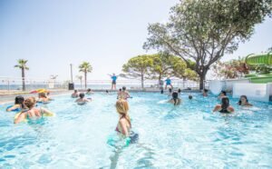 Personnes s'amusant dans une piscine lors d'une journée ensoleillée en Corse, entourées de palmiers et participant à des activités aquatiques dans un cadre convivial.