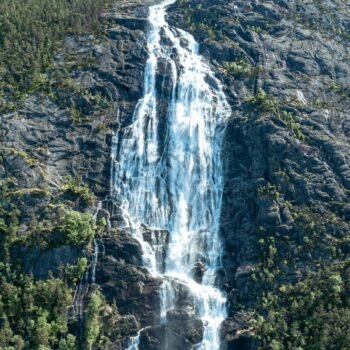 Cascade déferlante sur roche escarpée, entourée de verdure dense. Lumière solaire illuminant les gouttelettes en suspension.