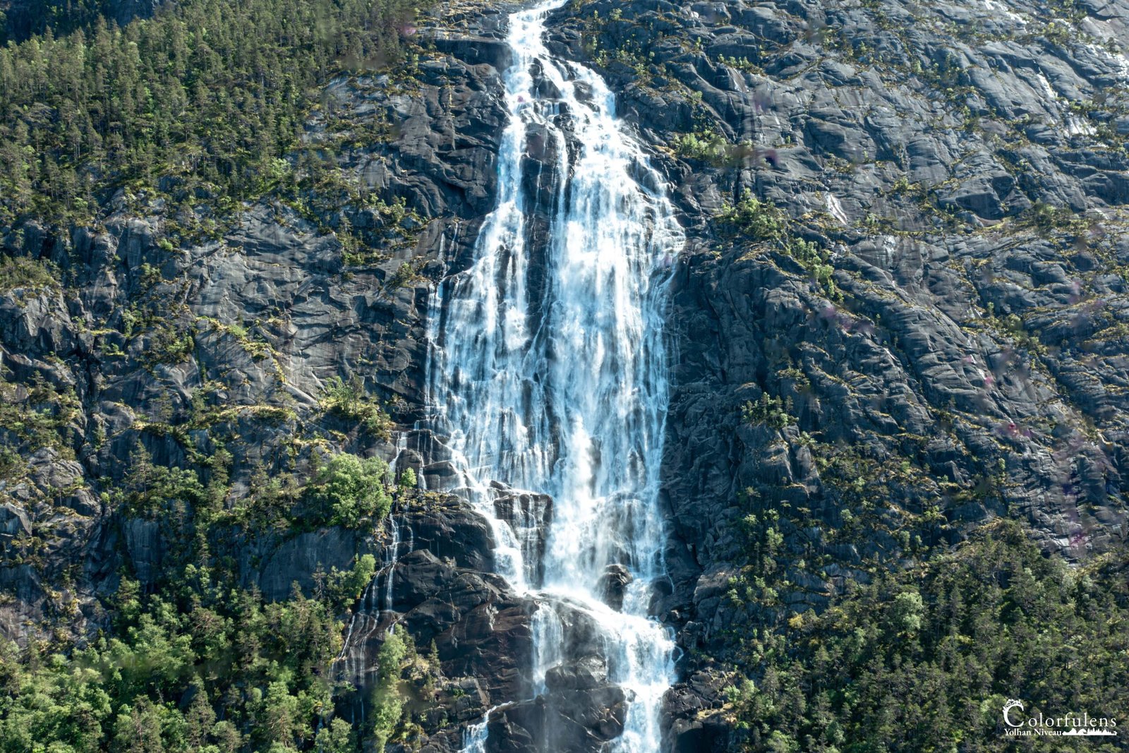 Cascade déferlante sur roche escarpée, entourée de verdure dense. Lumière solaire illuminant les gouttelettes en suspension.