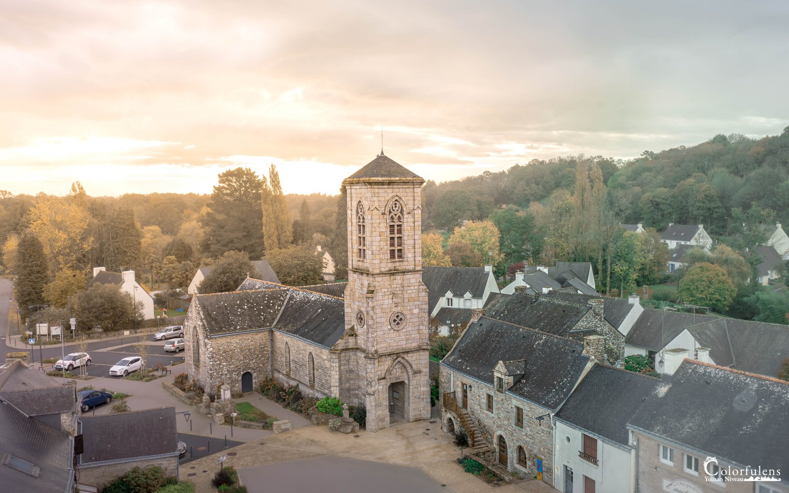 Vue aérienne d'une église au cœur d'un village français entouré de verdure et baigné de lumière au lever du soleil.