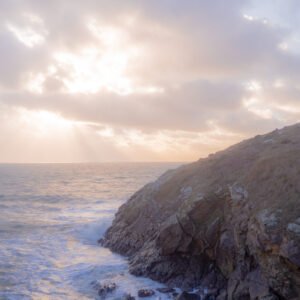Coucher de soleil spectaculaire illuminant une côte rocheuse avec des vagues percutantes, capturant la beauté de la nature contre un ciel nuageux.
