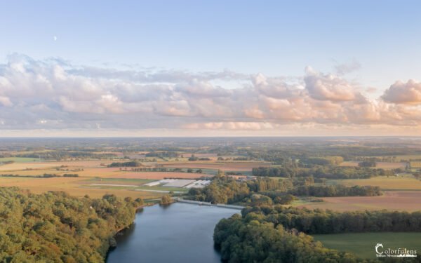 Vue aérienne d'un paysage de campagne au crépuscule avec un plan d'eau, des arbres et des nuages réfléchissant les dernières lueurs du jour.