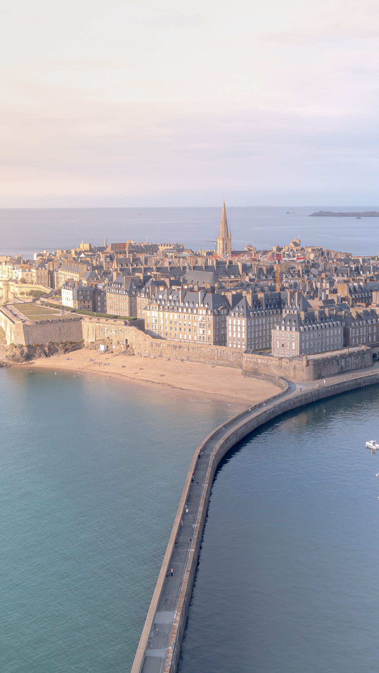 Vue aérienne de Saint-Malo sous une lumière matinale douce, révélant ses remparts historiques et ses voiliers au lever de soleil.