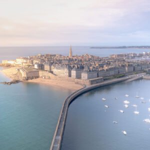 Vue aérienne de Saint-Malo sous une lumière matinale douce, révélant ses remparts historiques et ses voiliers au lever de soleil.