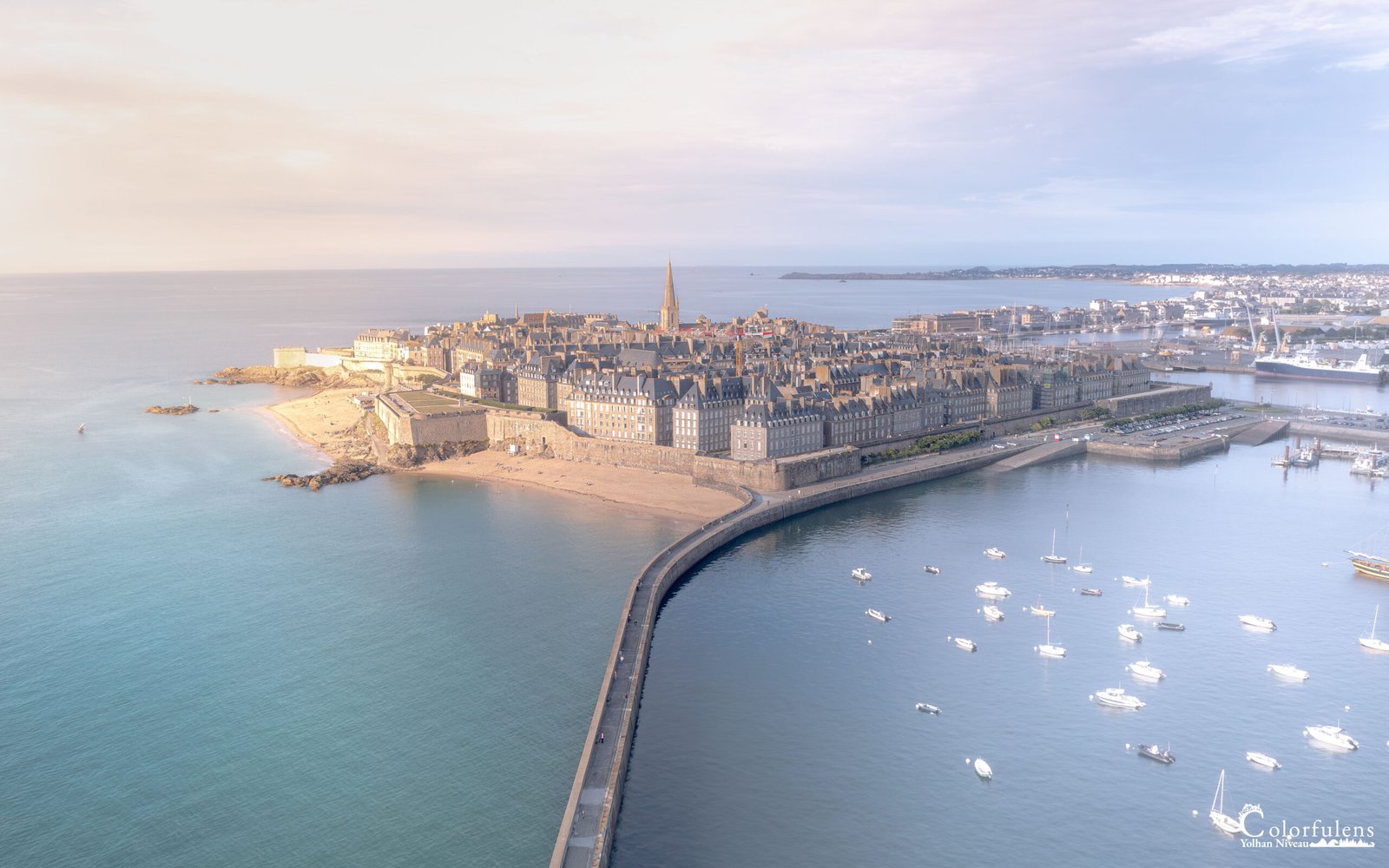 Vue aérienne de Saint-Malo sous une lumière matinale douce, révélant ses remparts historiques et ses voiliers au lever de soleil.