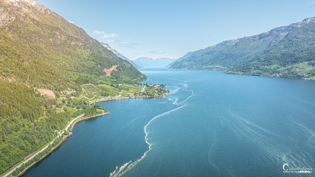 Vue aérienne du fjord de Hardanger en Norvège avec ses eaux bleues profondes, ses montagnes escarpées et ses quelques habitations au bord, reflétant la symbiose entre nature et civilisation.