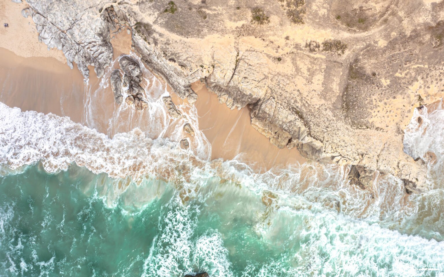 Vue aérienne d'une plage où le sable doré se mêle aux eaux turquoise et aux vagues éclatantes frappant des rochers majestueux, capturant la puissance et la beauté de la mer.