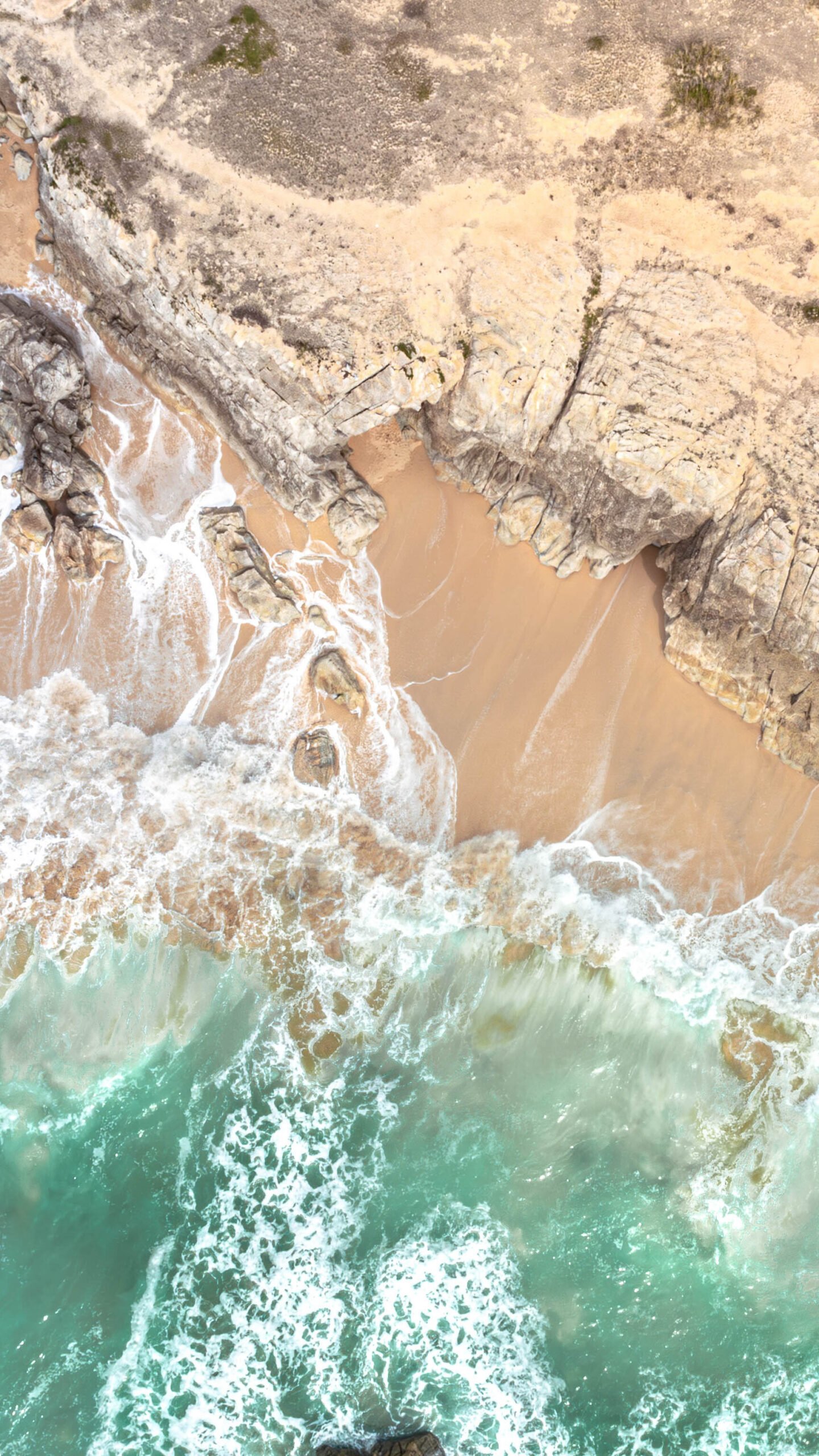 Vue aérienne d'une plage où le sable doré se mêle aux eaux turquoise et aux vagues éclatantes frappant des rochers majestueux, capturant la puissance et la beauté de la mer.