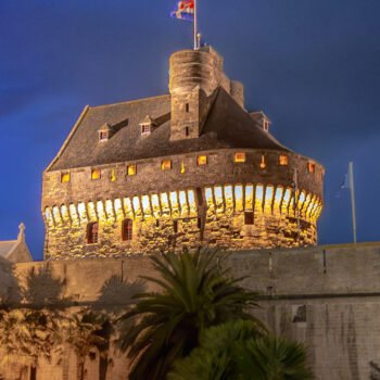 Forteresse majestueuse de Saint-Malo illuminée la nuit, avec des drapeaux flottant au sommet des remparts, représentant l'héritage historique de la ville bretonne.