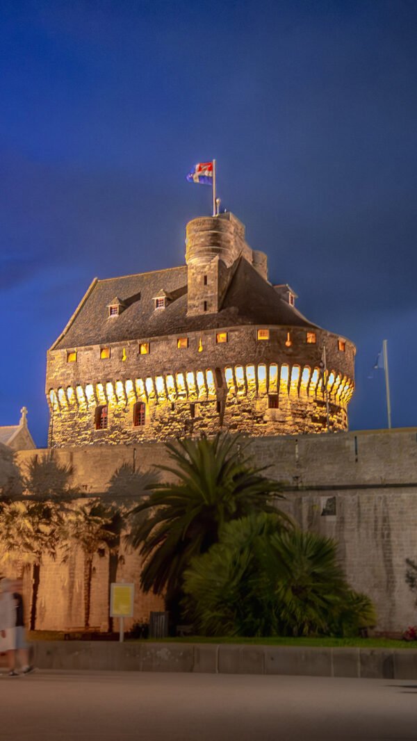 Forteresse majestueuse de Saint-Malo illuminée la nuit, avec des drapeaux flottant au sommet des remparts, représentant l'héritage historique de la ville bretonne.